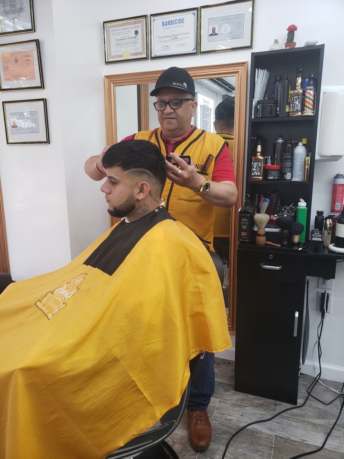 Daniel in his gold vest shaping a fade with a comb, framed licenses on the wall behind him